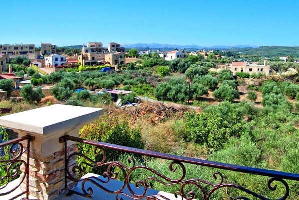 A balcony with a stone ledge and wrought iron railing overlooking a residential area with houses and olive trees.