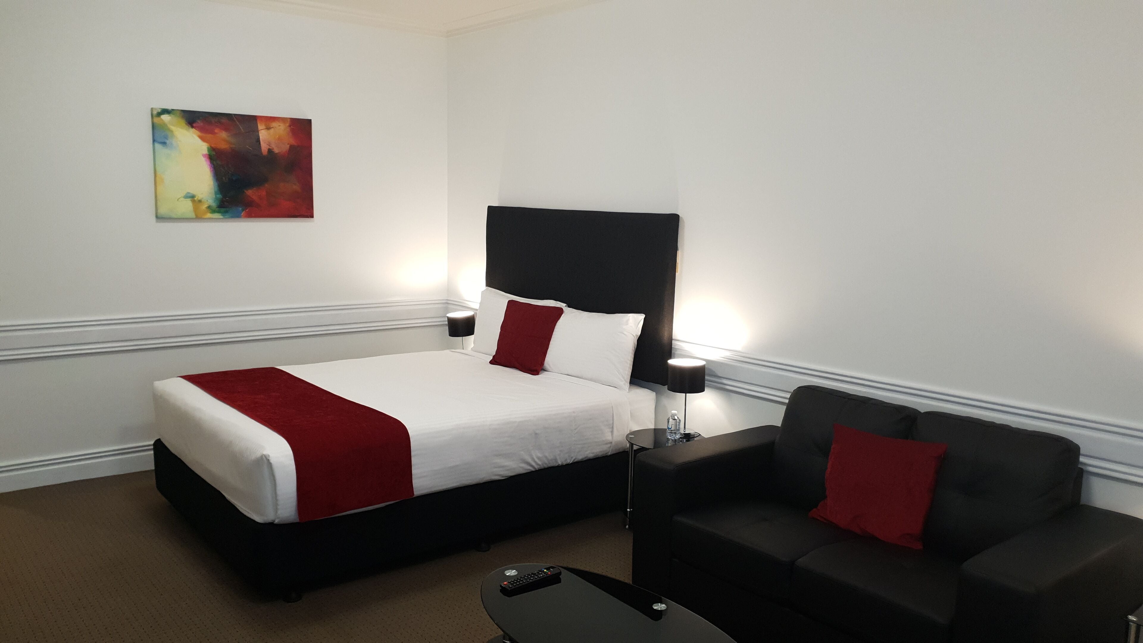 A hotel room with a bed, a black sofa, a small table with a remote control, and a painting on the wall.