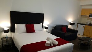 Ein Hotelzimmer mit einem großen Bett, einer roten Tagesdecke, zwei Kissen, einem Sessel, einem kleinen Tisch und einer Küchenzeile im Hintergrund.