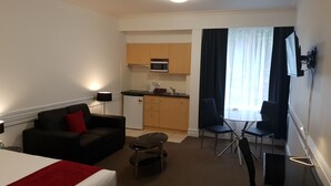 Ein kompaktes Hotelzimmer mit Bett, einer kleinen Sitzecke, einem Couchtisch, einem Schreibtisch, einem Sessel sowie einer Küchenzeile mit Mikrowelle und einem an der Wand befestigten Fernseher.