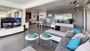A spacious living area with a sofa, two round tables, a television displaying a movie, and a kitchen in the background. - Villa SEA VIEW, 5' from Grand Case Beach, private pool, huge privacy (saint martin)