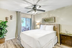 A hotel room with a bed, a ceiling fan, a nightstand, a lamp, a mirror, and a potted plant.