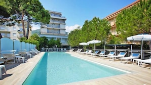 A rectangular swimming pool with lounge chairs and umbrellas along the sides, surrounded by trees and buildings. - Apartment in Pietra Ligure with garden (Pietra Ligure)