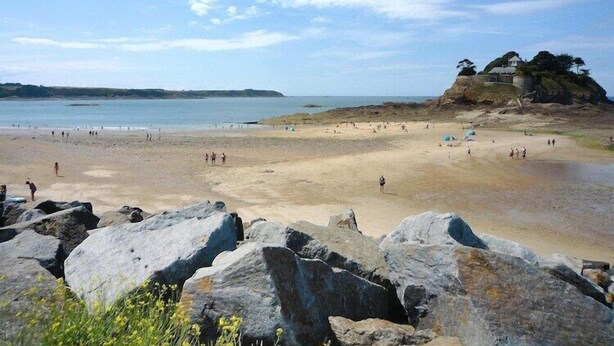 Een strand met grote rotsen op de voorgrond, een zandstrand en een kleine heuvel waarop een gebouw staat.