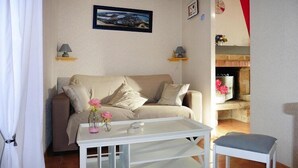 Een woonkamer met een beige bank, een witte bijzettafel met de tekst 'JOURNAUX' en een kleine witte stoel. Er is een open haard met roze bloemen op de schoorsteenmantel.