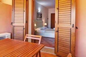 A room with a bed, a table, and a chair. There is a door with a wooden shutter, and a picture on the wall.