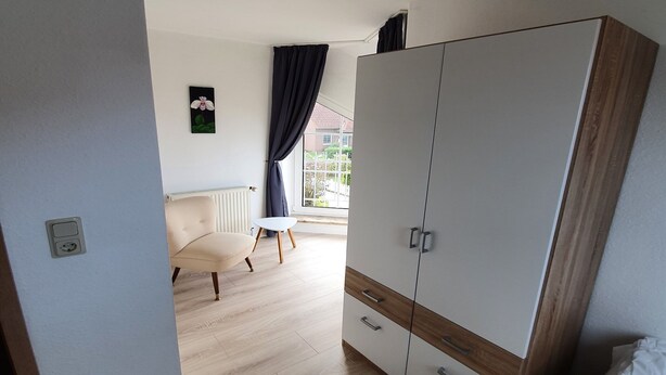 Ein Schlafzimmer mit großem Kleiderschrank, einem Bett, einem Sessel und einem Fenster mit Vorhängen.