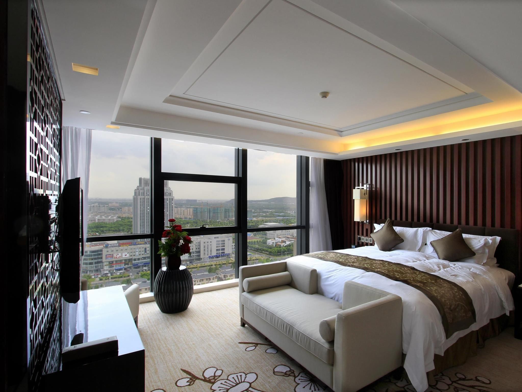 A modern hotel room with a large bed, a sofa, a TV, and a view of the cityscape.