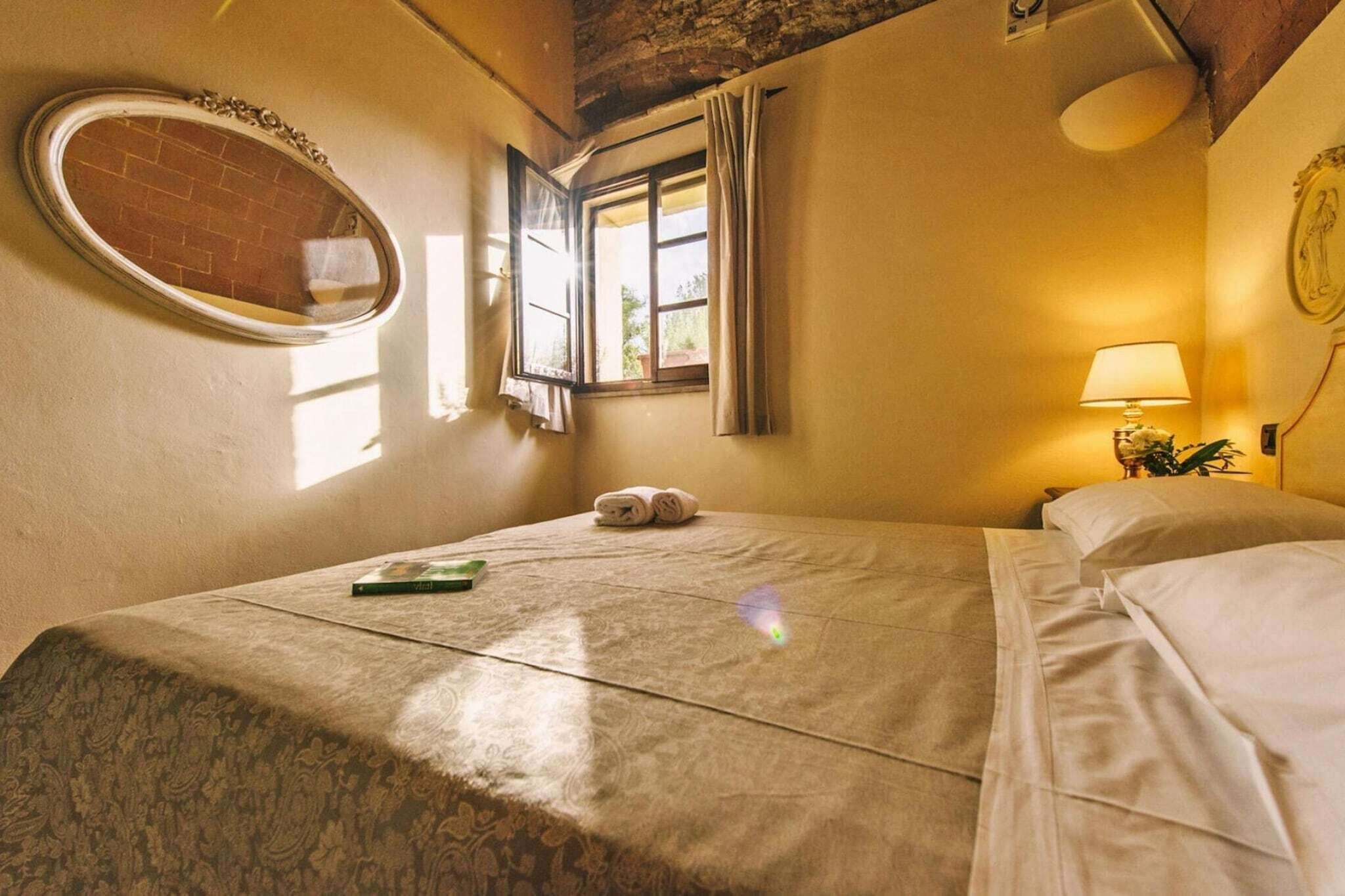 Ein Hotelzimmer mit einem Bett, einem runden Spiegel, einem Fenster mit Vorhängen, einer Nachttischlampe und einem kleinen Tisch mit einer Pflanze.