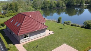 A two-story building with a red roof, situated next to a calm lake with trees and wind turbines in the background. - Appartementhaus am Kiessee, Westerholt (Westerholt)