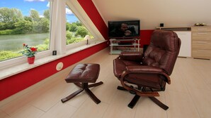 A modern living room with a large window, a leather chair, a TV, and a small table. - Appartementhaus am Kiessee, Westerholt (Westerholt)