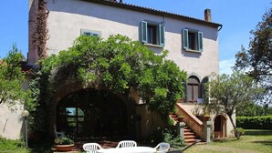 Una casa de dos pisos con un porche cubierto, una escalera que conduce al nivel superior y un jardín con mesa y sillas.