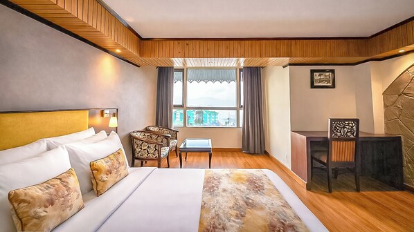 Habitación de hotel con cama, escritorio, silla y vistas a un paisaje urbano a través de amplias ventanas.