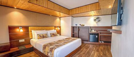 A hotel room with a wooden ceiling, a bed with a patterned bedspread, a small desk with a computer, and a wall-mounted fan.