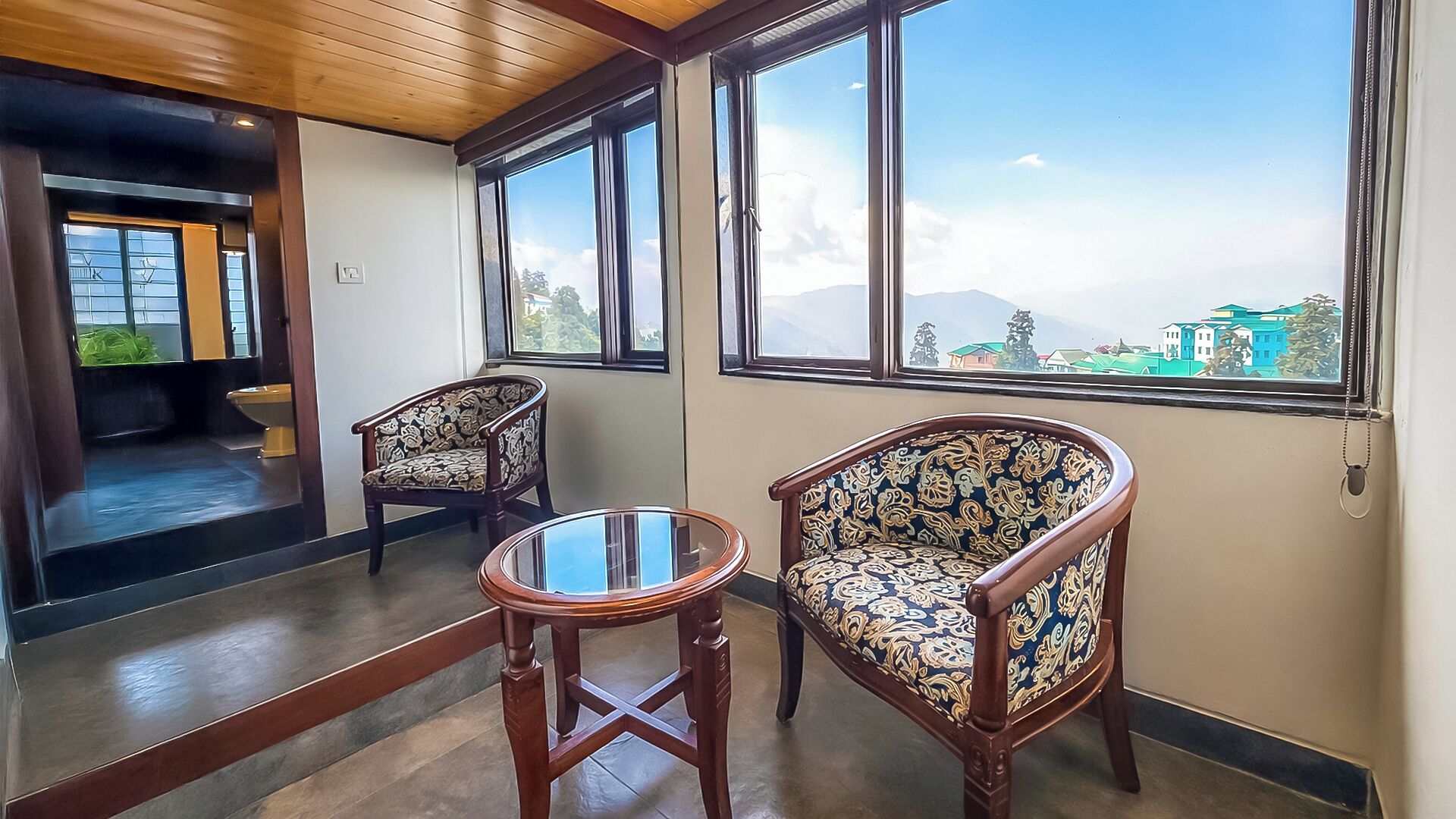 A room with two ornate chairs, a small round table, and large windows offering a mountain view.