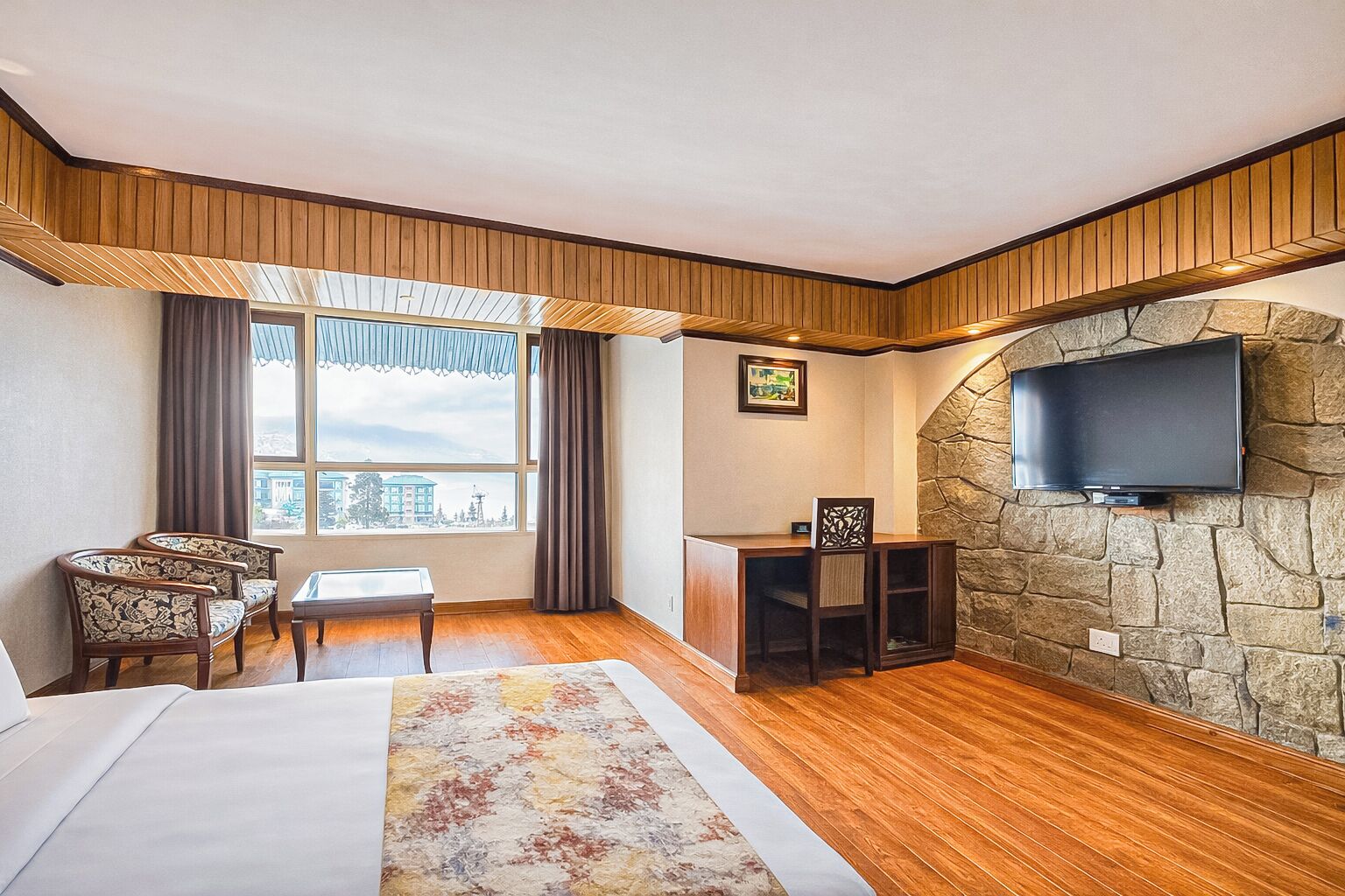 A hotel room with a bed, a desk, a chair, a TV mounted on a stone wall, and a large window with curtains.