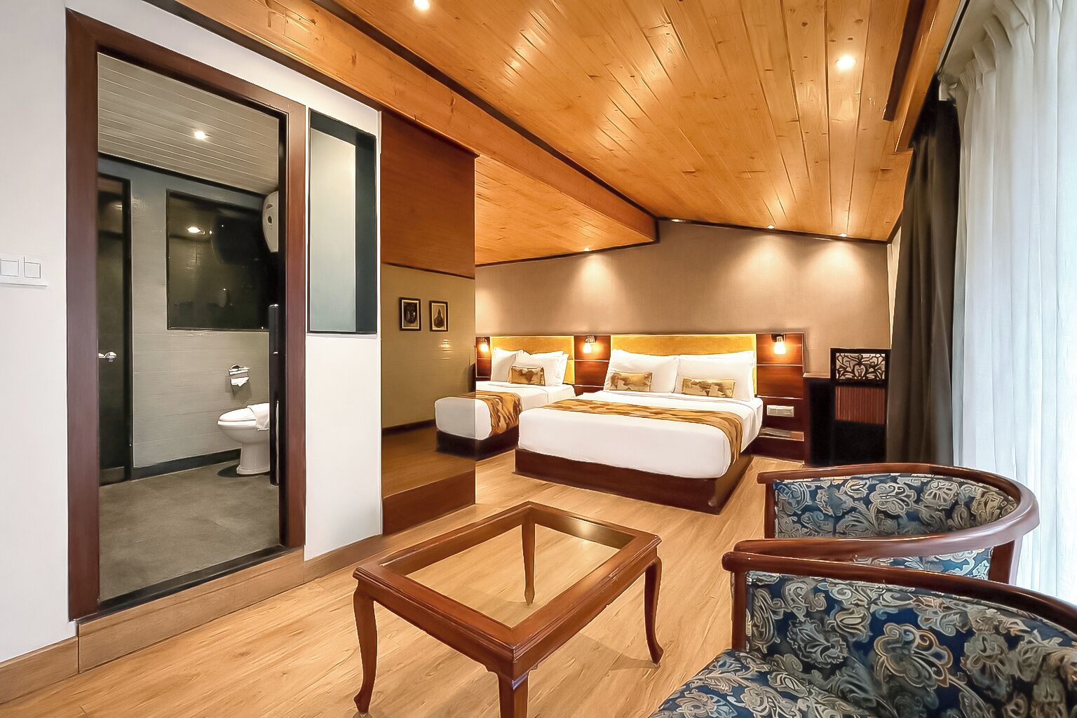 A hotel room with a wooden ceiling, a large bed with two bedside tables, a sofa, and a glass door leading to a bathroom.