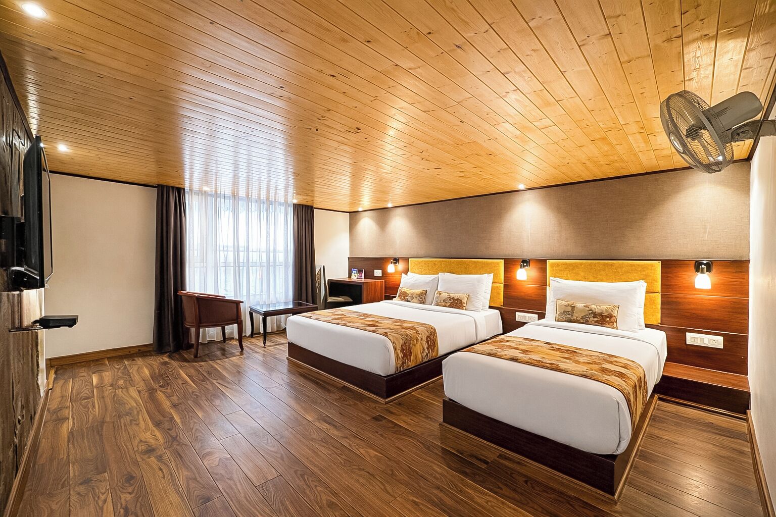 A hotel room with two beds, wooden flooring, a desk, and a TV.