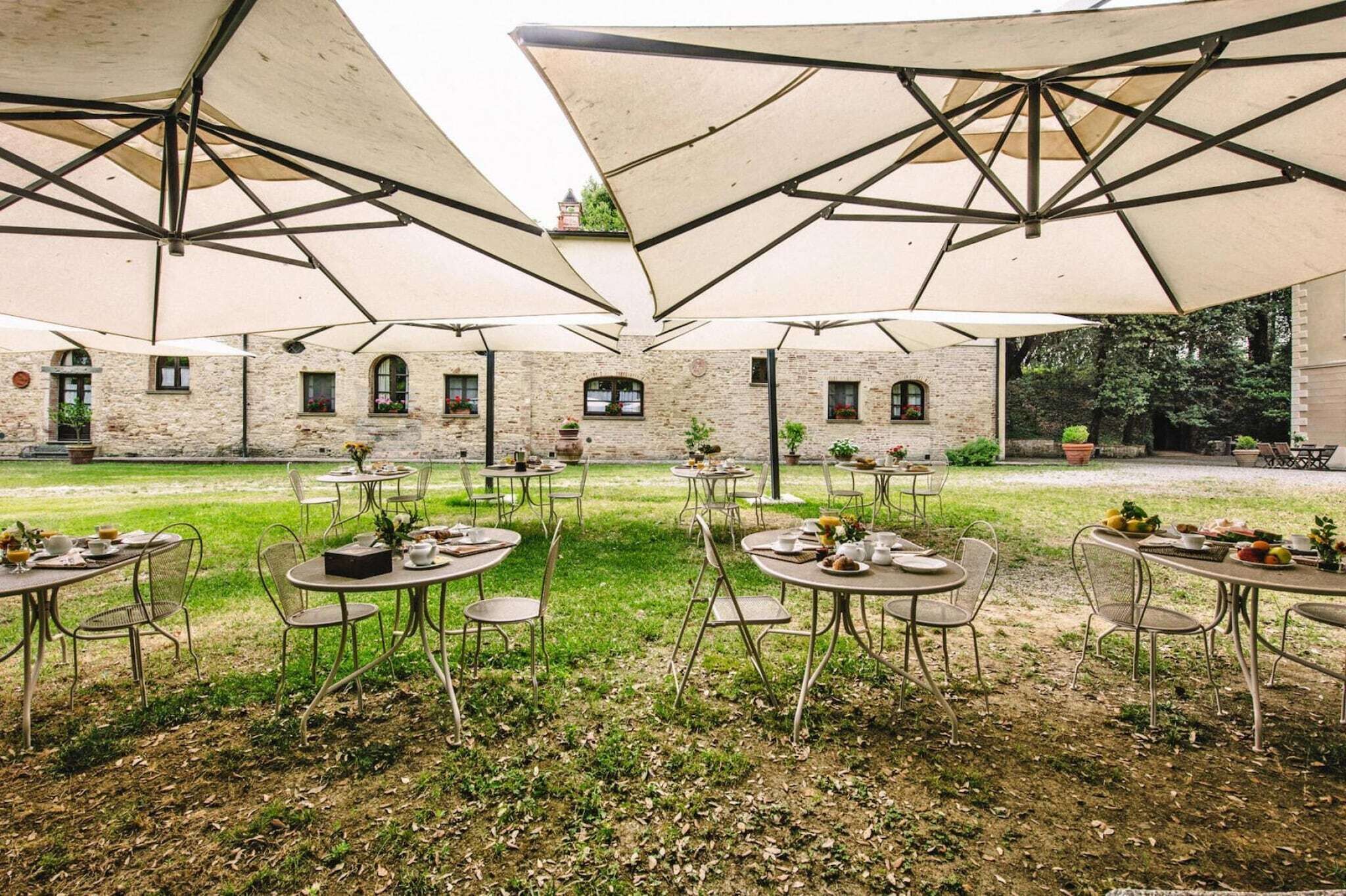 Un área de comedor al aire libre con mesas y sillas bajo grandes sombrillas, rodeada de edificios de piedra.