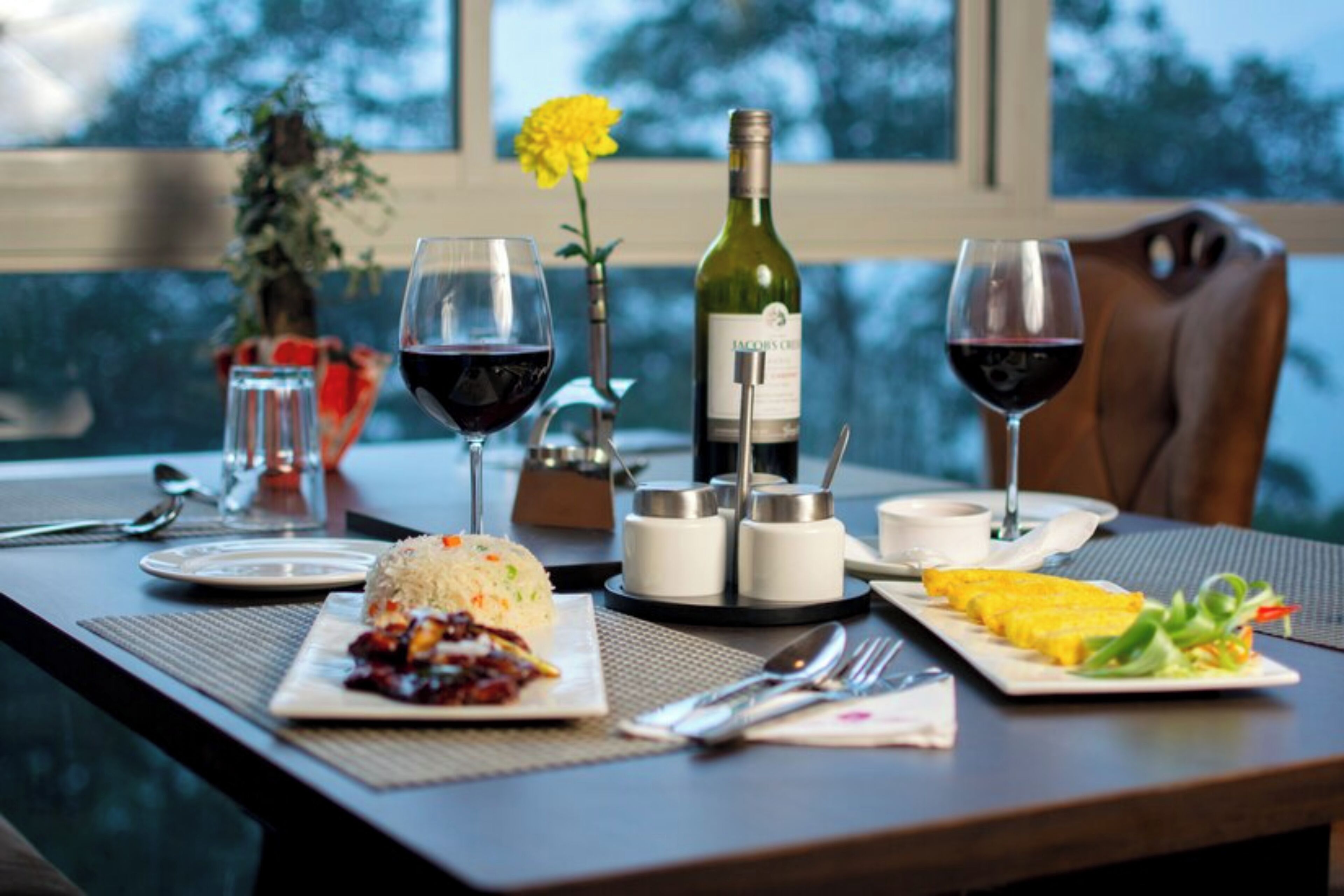 A dining table with a bottle of wine, two wine glasses, a plate of food, and a flower.