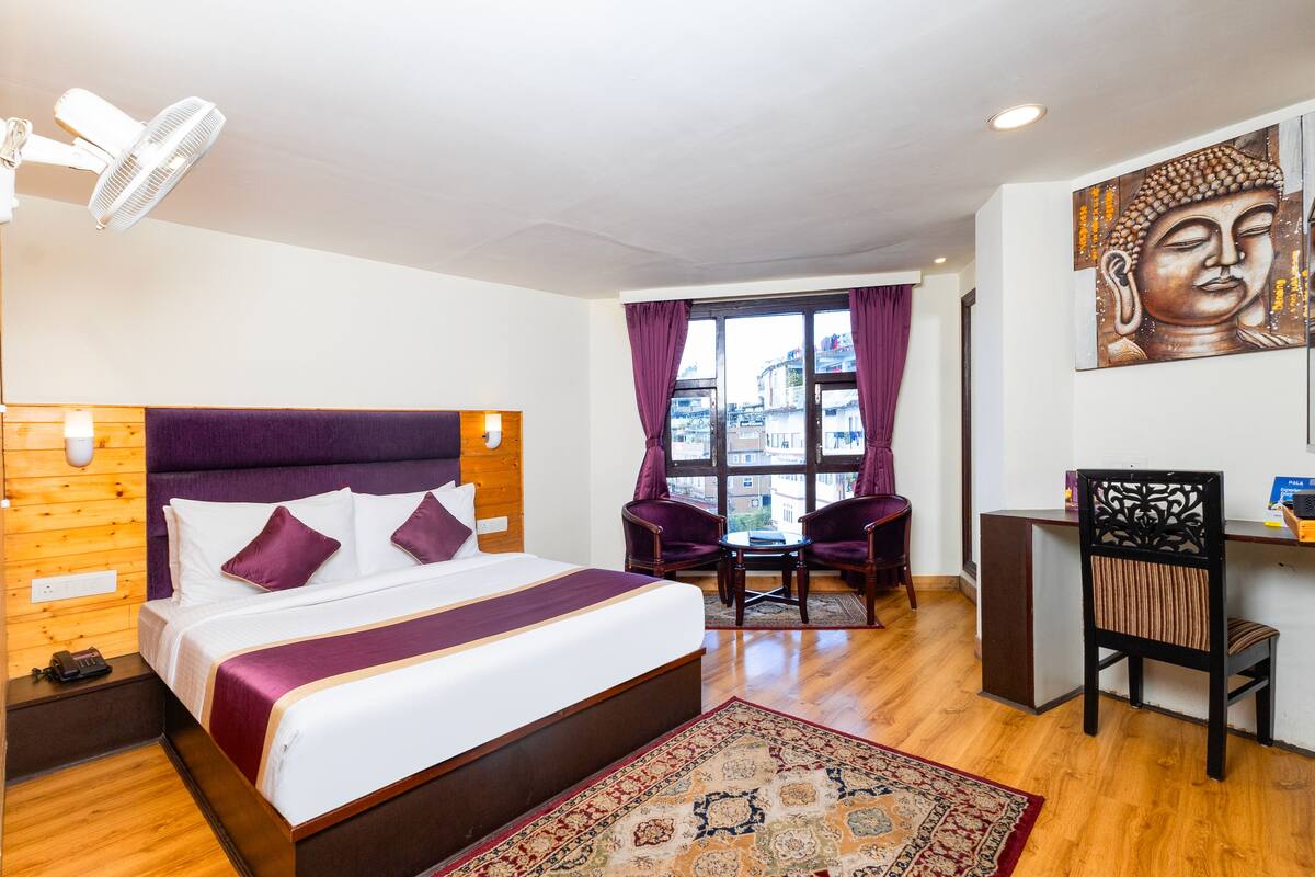 A hotel room with a large bed, a desk, two chairs, a window with curtains, a Buddha painting, and a fan.
