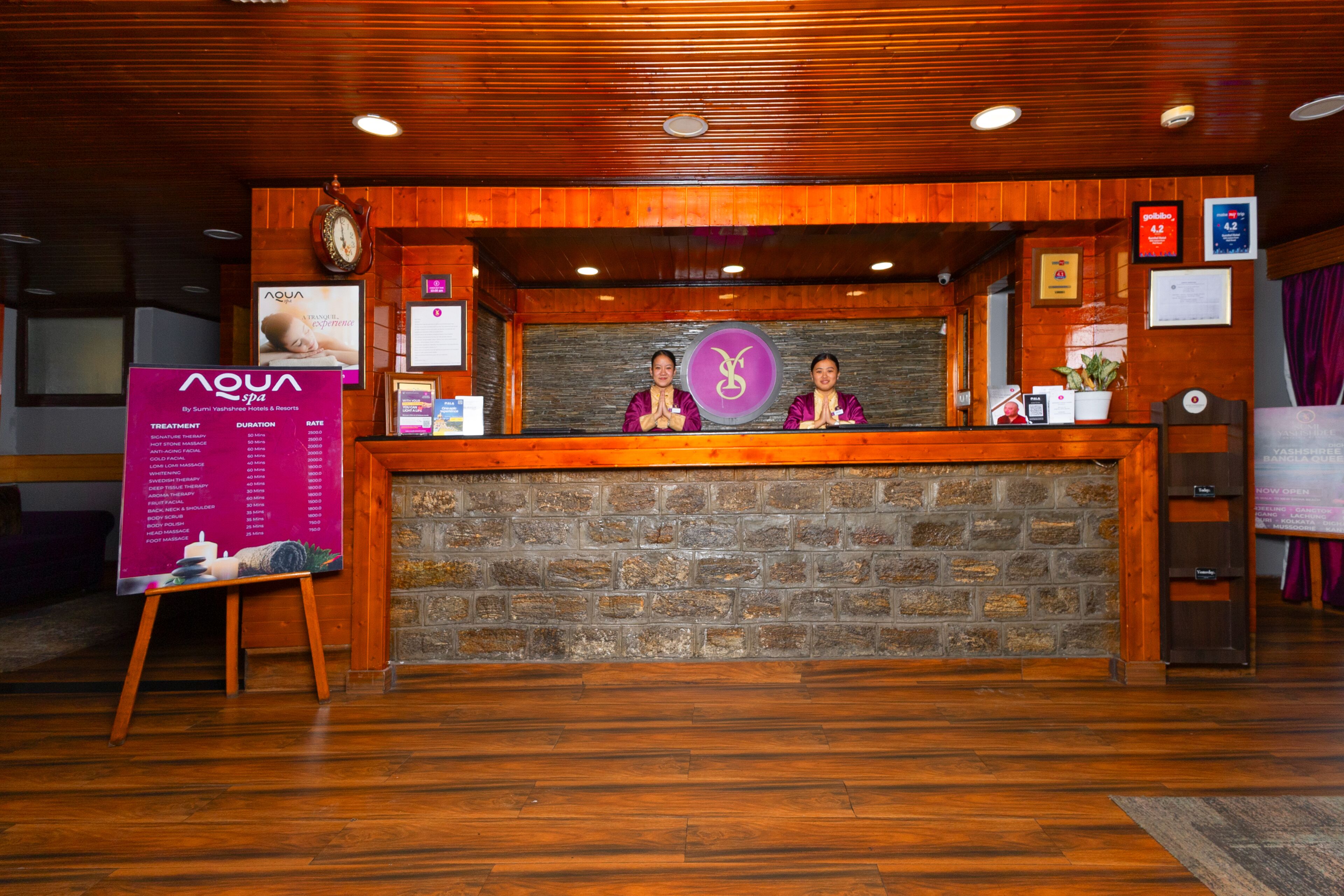 A reception desk with a wooden finish and a brick wall backdrop. Two receptionists are seated behind the desk, which has a logo and a clock above it.