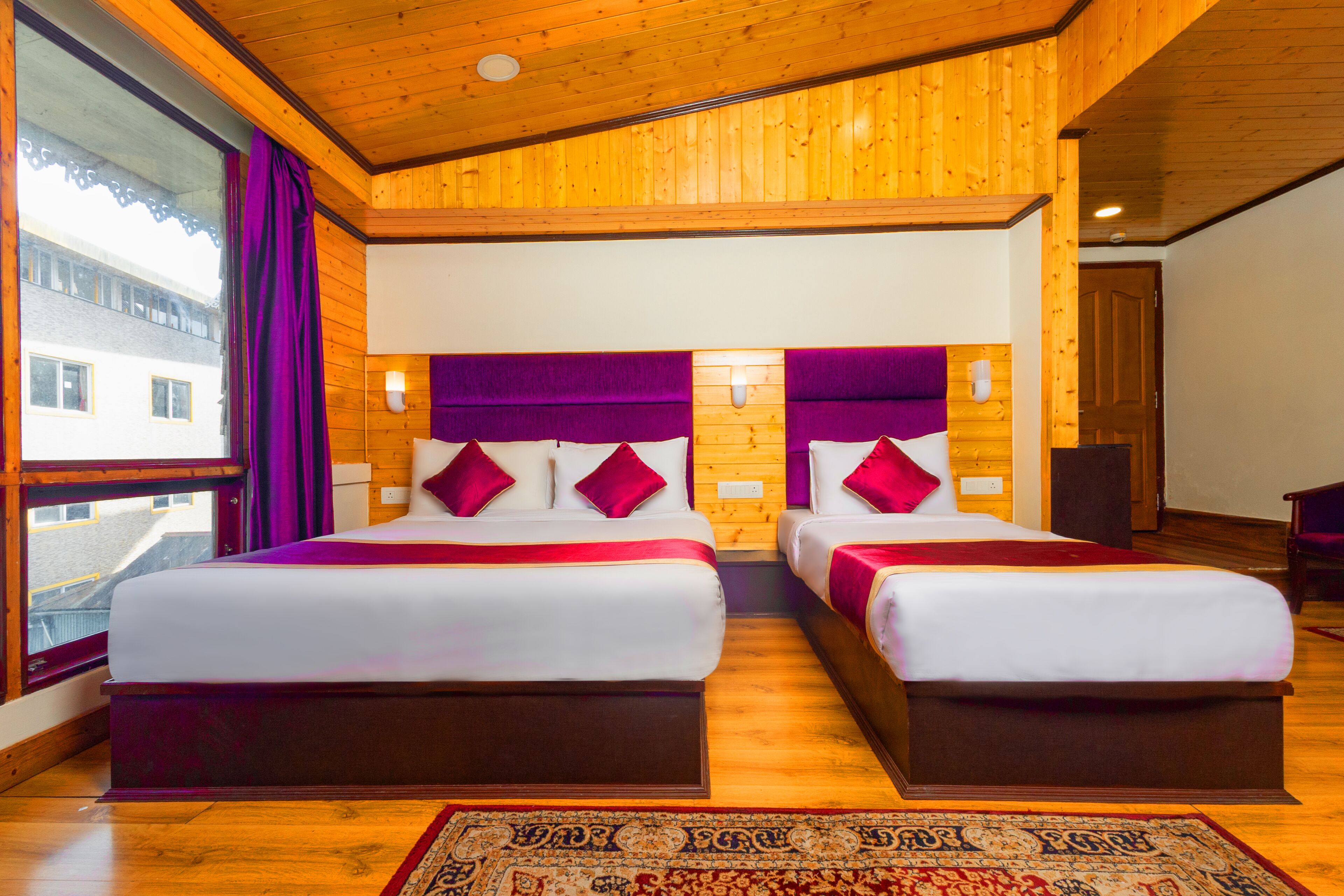 Ein Doppelzimmer mit zwei Betten, Holzboden, großen Fenstern mit violetten Vorhängen und einem gemusterten Teppich.