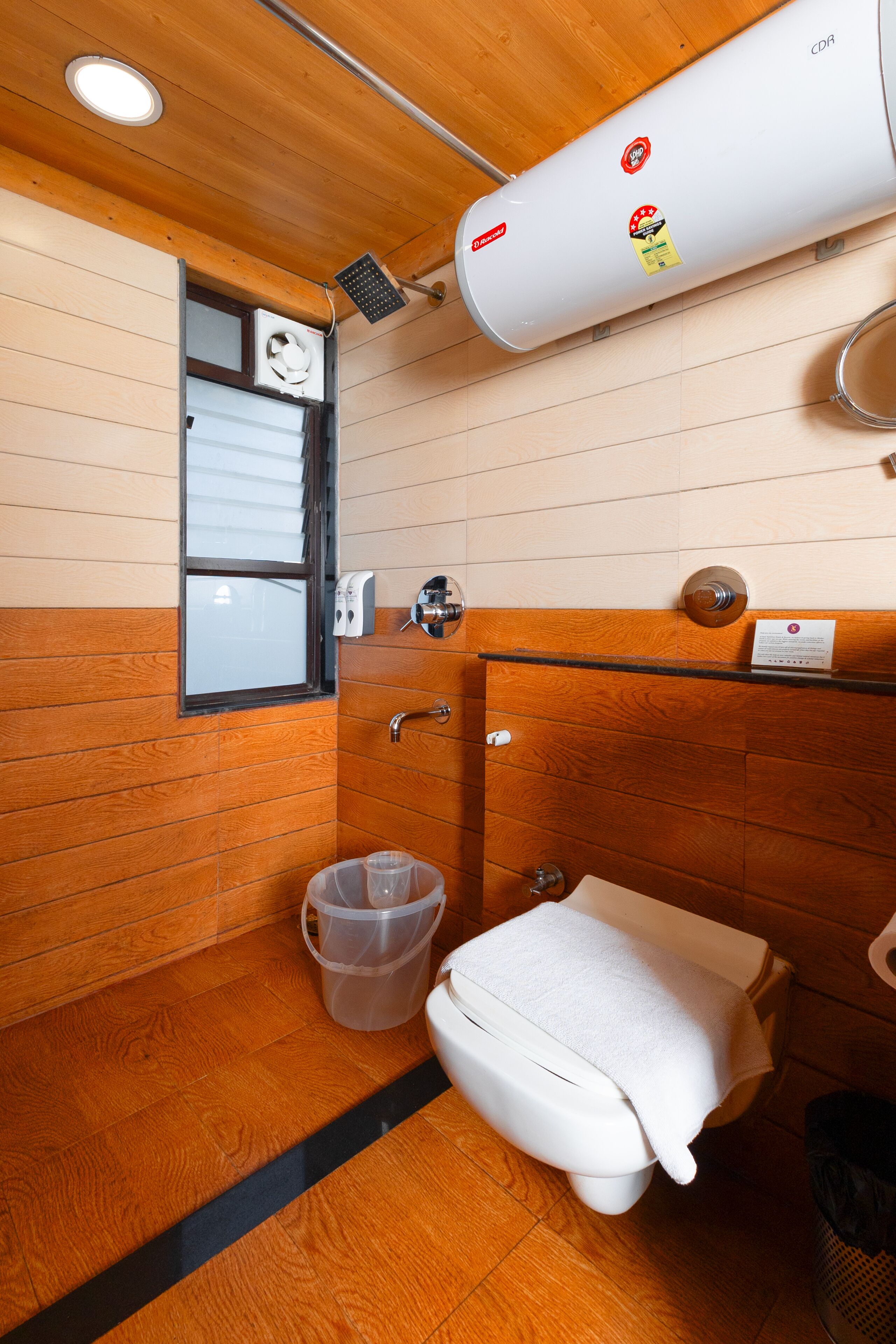 Une petite salle de bain avec un lambris en bois, des toilettes blanches, un espace douche et une petite fenêtre.