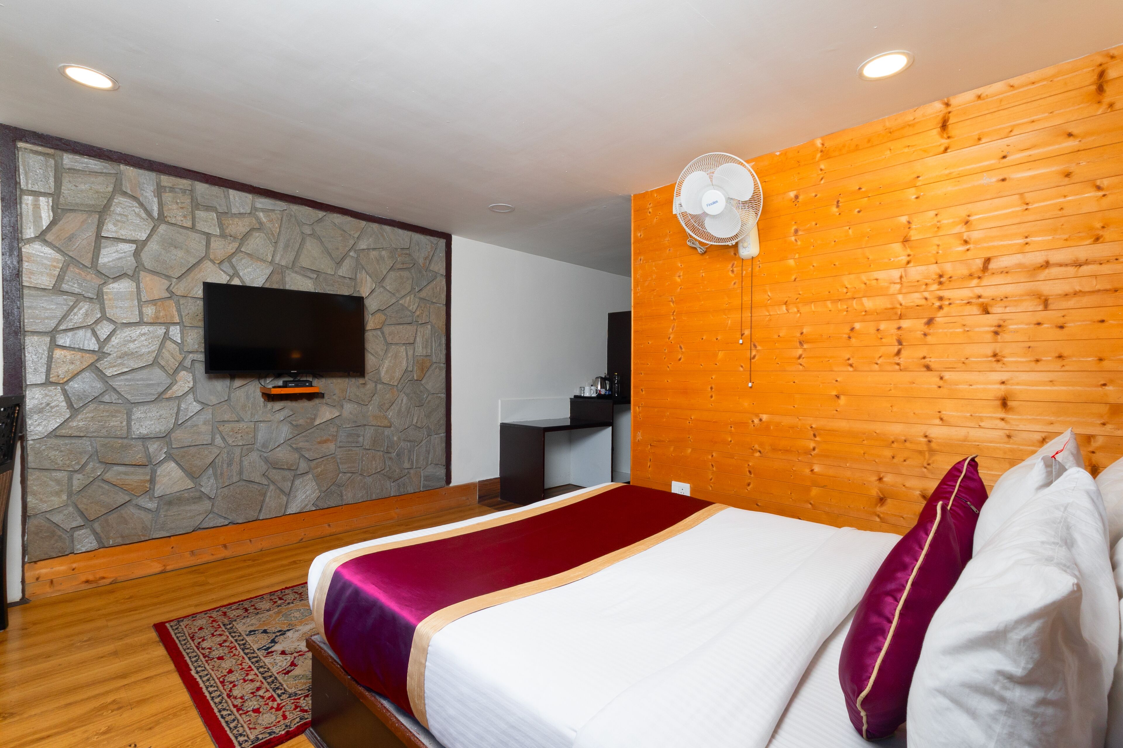 Een hotelkamer met een stenen muur, houten lambrisering, een bed met wit en paars beddengoed, een aan de stenen muur bevestigde televisie en een plafondventilator.