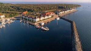 Vista aérea de un pueblo costero con un puerto deportivo, casas y una colina boscosa al fondo.