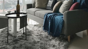 A modern living room with a grey sofa, a small round table with a vase and glass, and a fluffy grey rug. - Boulevard Service Apartment KL by Airhost (Kuala Lumpur)