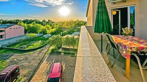 A balcony with a table and chairs, overlooking a garden and a road with parked cars.