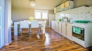 A modern kitchen with wooden flooring, white cabinetry, a dining area with a white table and chairs, and a window providing natural light.