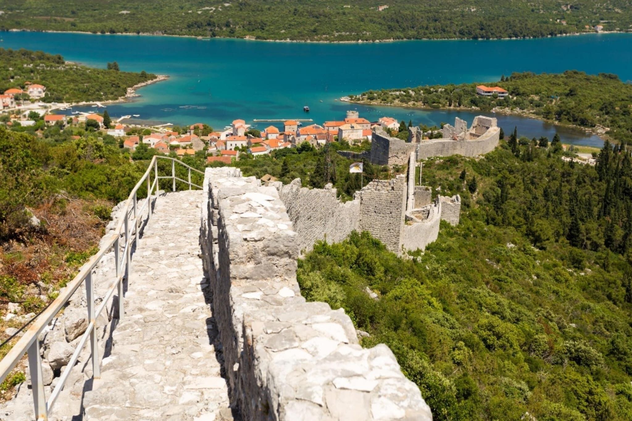 A stone pathway leading to a historic stone wall with a view of a coastal town and a large body of water.