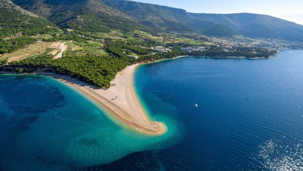 An aerial view of a coastal area with a beach, clear waters, and mountainous terrain.