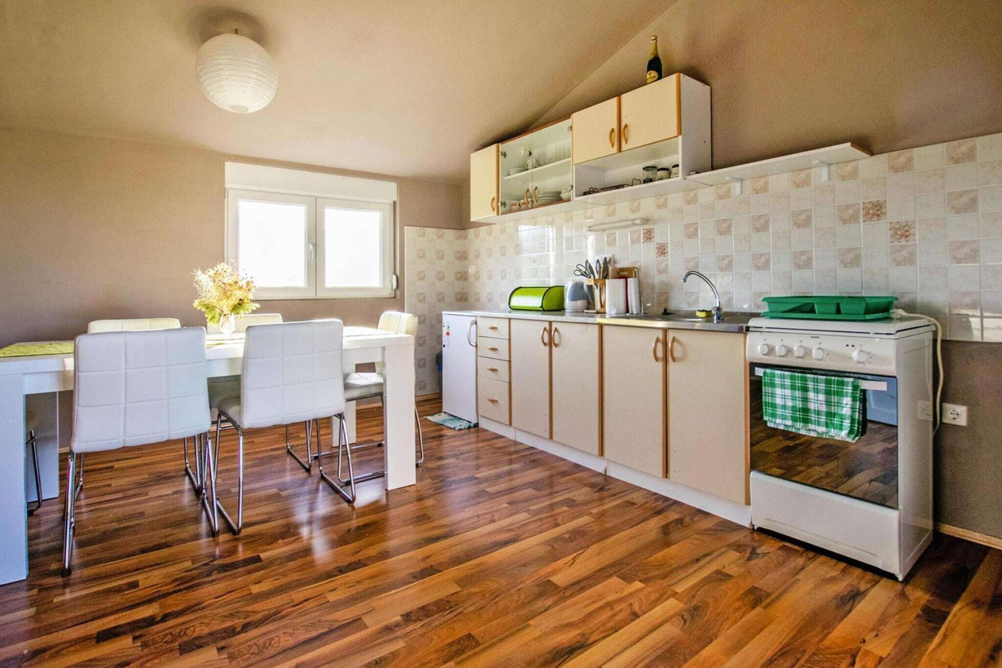 Une cuisine avec un plancher en bois, des armoires blanches, un four encastré et un coin repas comprenant une table et des chaises blanches.