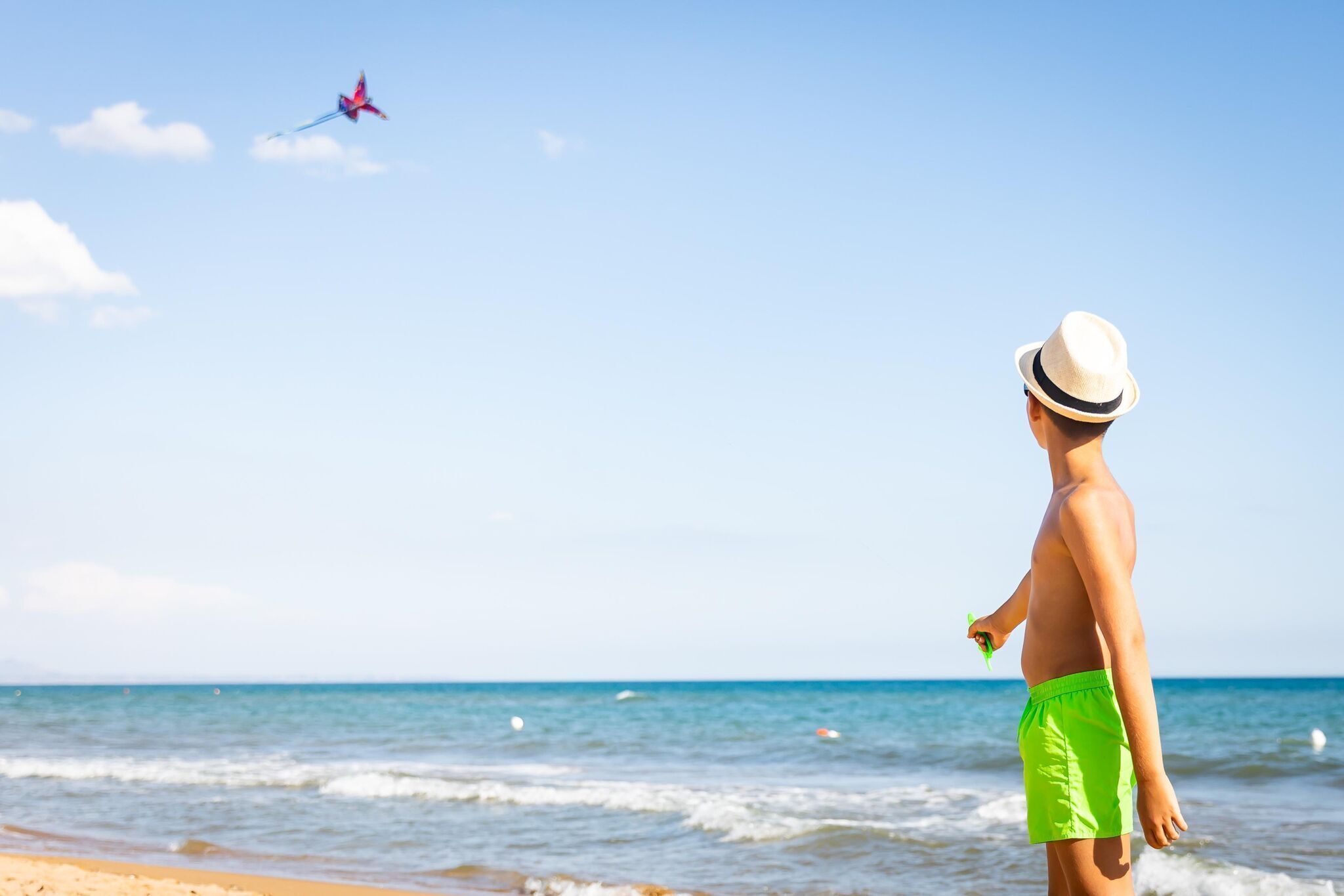 A beach scene with a person in green swim trunks and a hat, holding a kite.