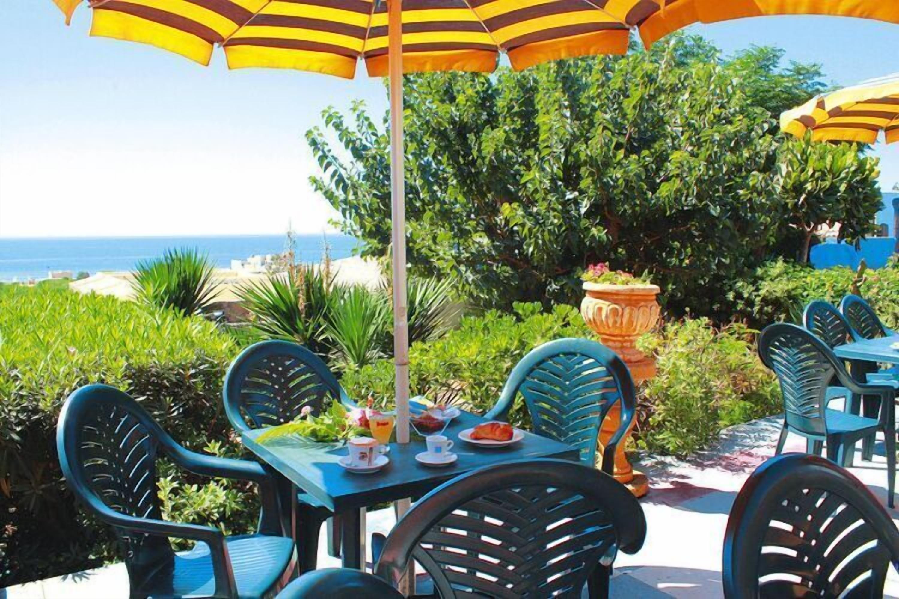 Un espace extérieur aménagé pour les repas, avec des tables et des chaises, un grand parasol et une vue sur l’océan.