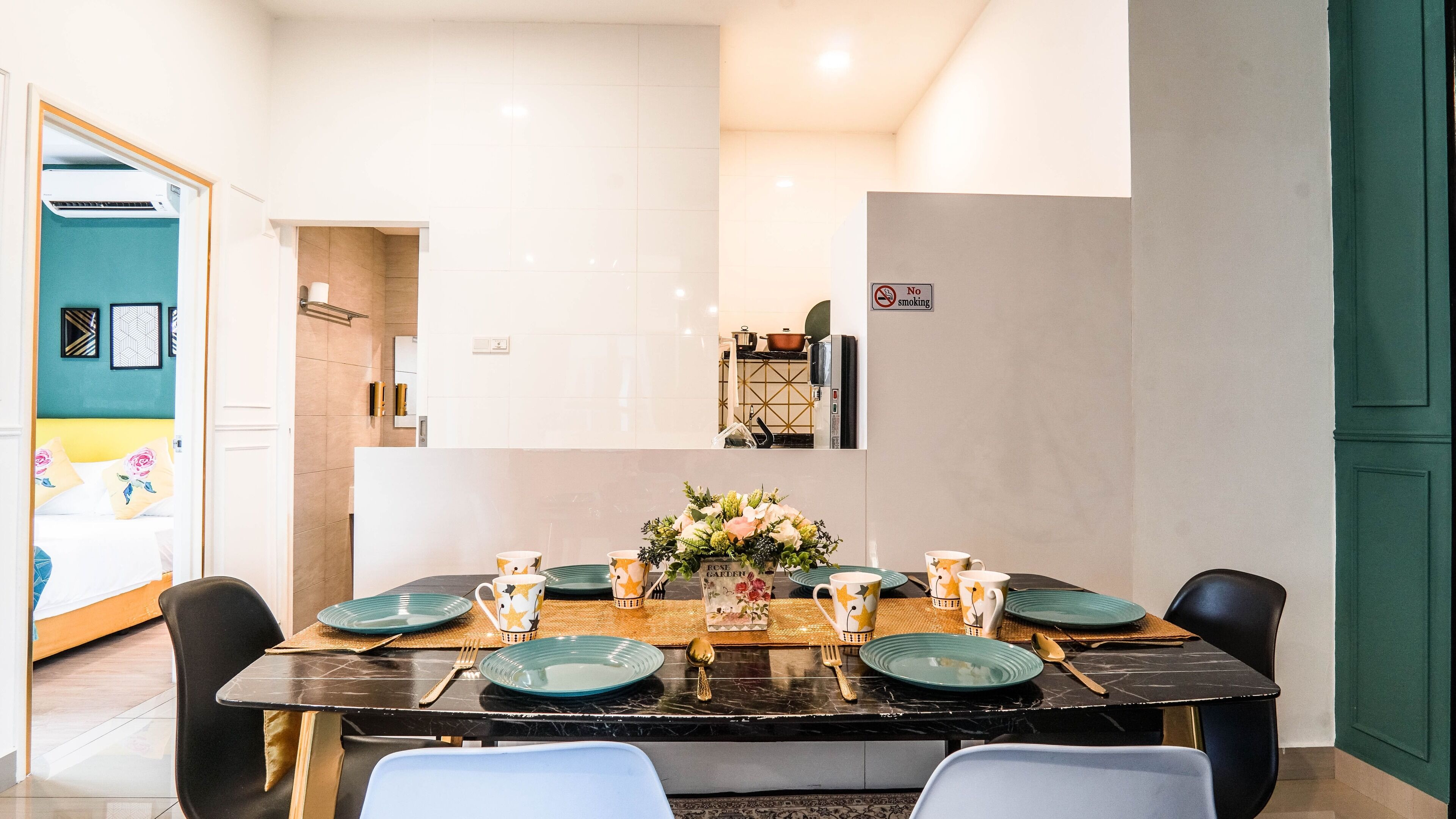A dining area with a table set for four, a kitchen in the background, and a view of a bedroom with a bed and a bathroom.
