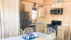 Una cocina compacta con armarios de madera, un refrigerador de acero inoxidable, un microondas y una estufa. Hay una mesa de comedor con sillas y una bandeja con tazas y platos.