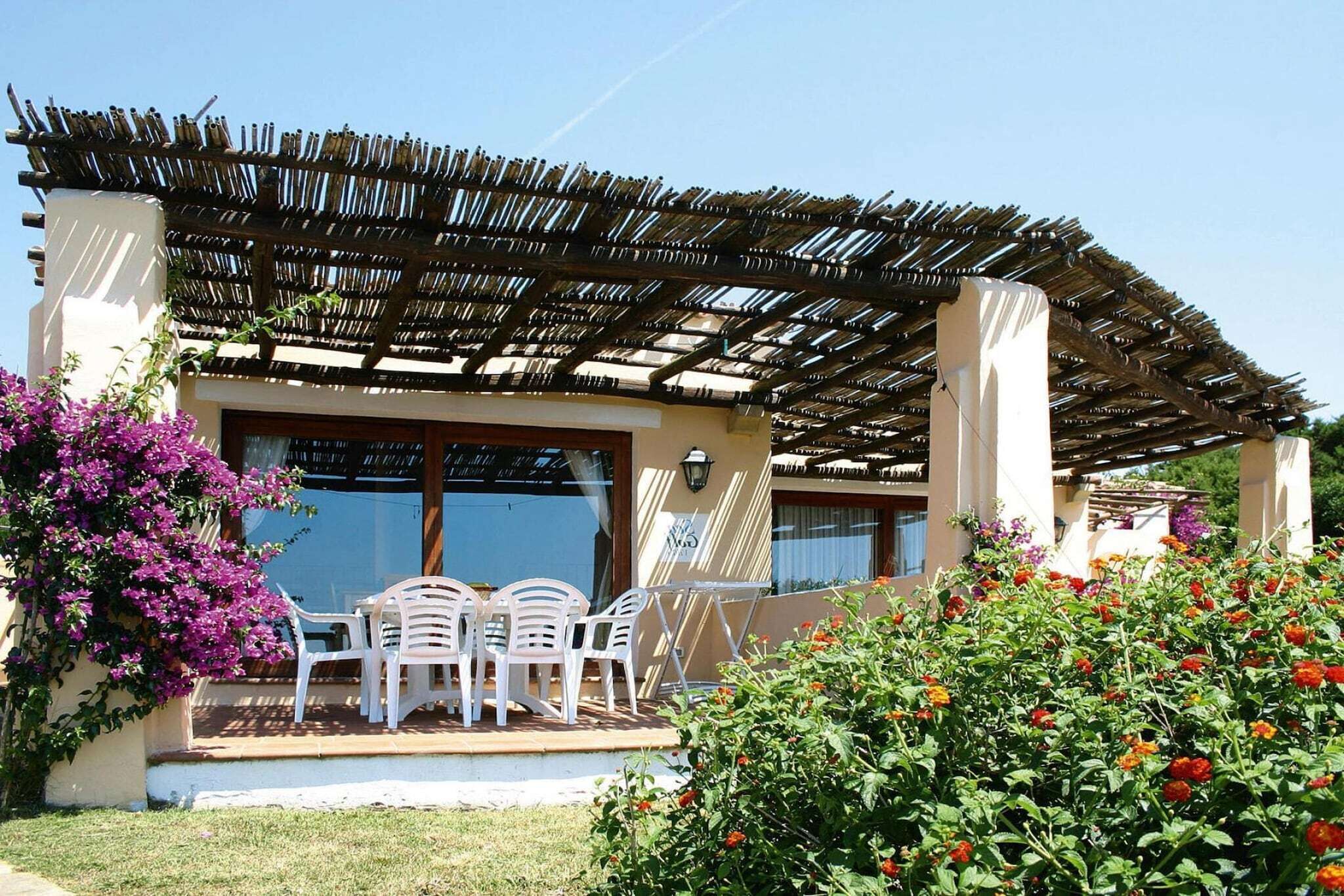 Et overdækket udendørs siddeområde med pergola, et bord med stole og farverige blomster.