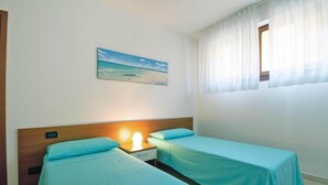 Une chambre avec deux lits, une table de chevet avec une lampe, un tableau représentant une scène de plage et une fenêtre avec des rideaux légers.