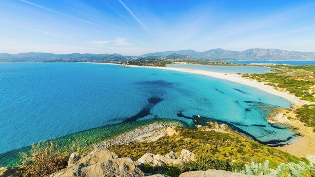 Un paysage côtier avec une eau d’un bleu limpide, une plage de sable et des montagnes à l’horizon.