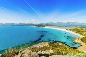 Un paysage côtier avec une eau d’un bleu limpide, une plage de sable et des montagnes à l’horizon.