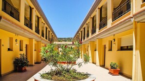 Une cour bordée de bâtiments jaunes, agrémentée de plantes en pot et surplombée d’un ciel bleu clair.