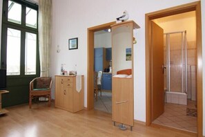 Ein Zimmer mit Holzboden, einem Sessel, einem Schrank, einer zur Toilette führenden Tür und einem Fenster mit Vorhängen.
