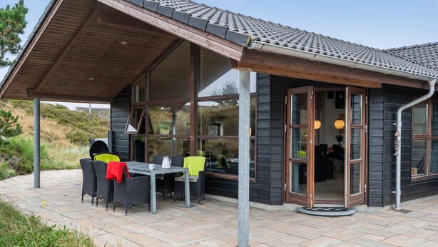 Un espace extérieur abrité, aménagé pour les repas, avec une table et des chaises, de grandes fenêtres et une porte donnant sur l’intérieur.