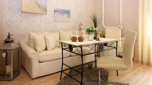 Un salon chaleureux avec un canapé beige, une table basse en verre et un fauteuil blanc. Deux tableaux ornent le mur, ainsi qu’un miroir décoratif et une petite plante posée sur la table.