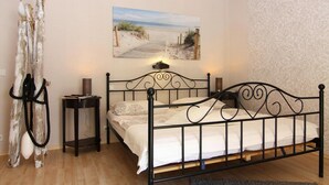 Une chambre avec un lit en métal, une table de chevet avec une lampe, une petite table ronde sur laquelle repose un livre, et une décoration murale sur le thème de la plage.