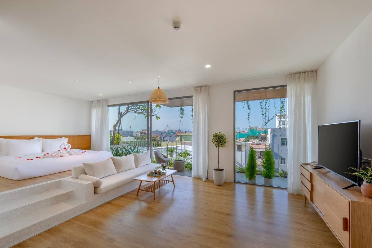 A modern hotel room with a large bed, a sofa, a TV, and a balcony with a view of the city.